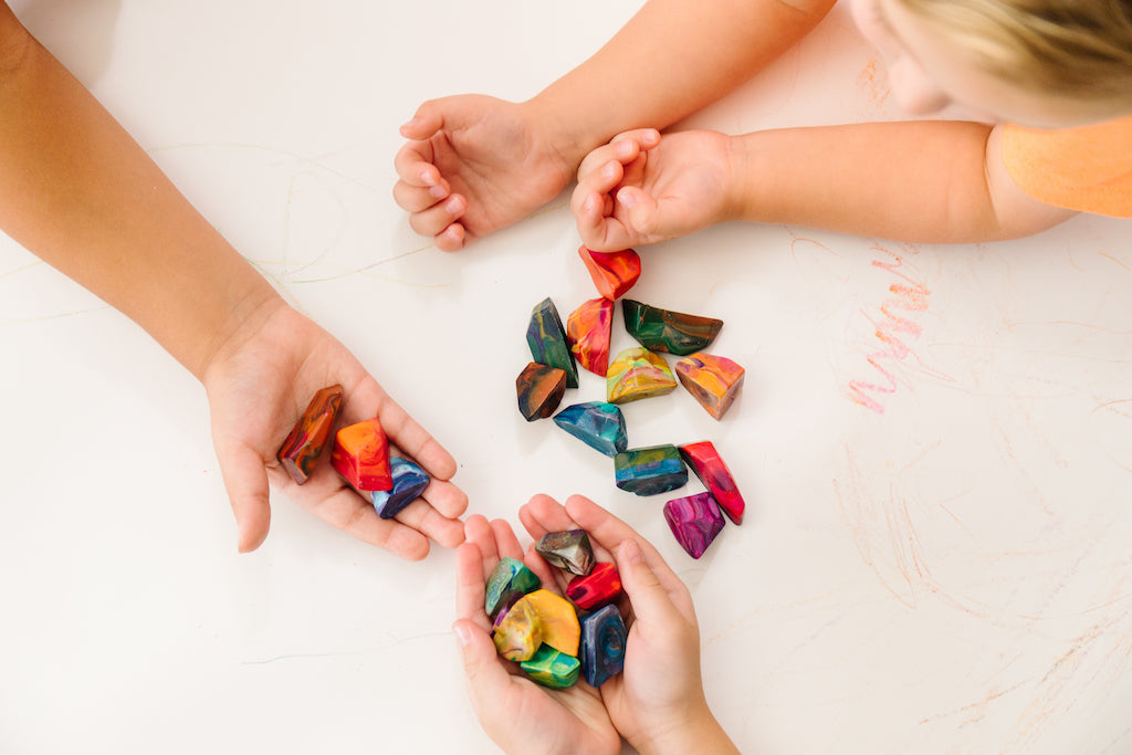 Two children arms and hands shown on a white background (paper). An older child holds 9 multicolored rock crayons in their hands while the other colors with 11 rock crayons spread onto of the white background. A red scribble is shown on the white paper.