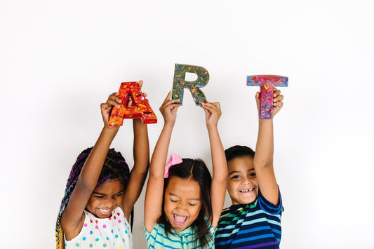 Three kids holding oversized rainbow crayons spelling the word ART. Gift guide article for clear the list, teacher wish lists on Amazon. 