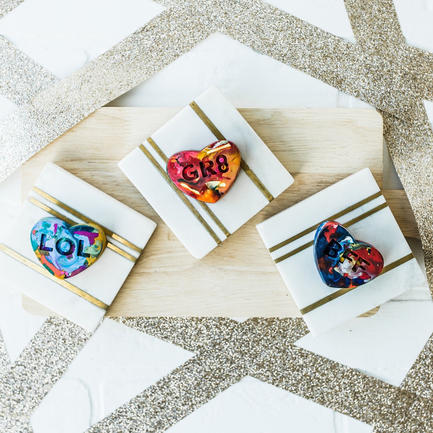 Three multi-colored conversation heart valentine crayons for kids are displayed on a white and gold background with a wooden charcuterie board as a photo prop. 