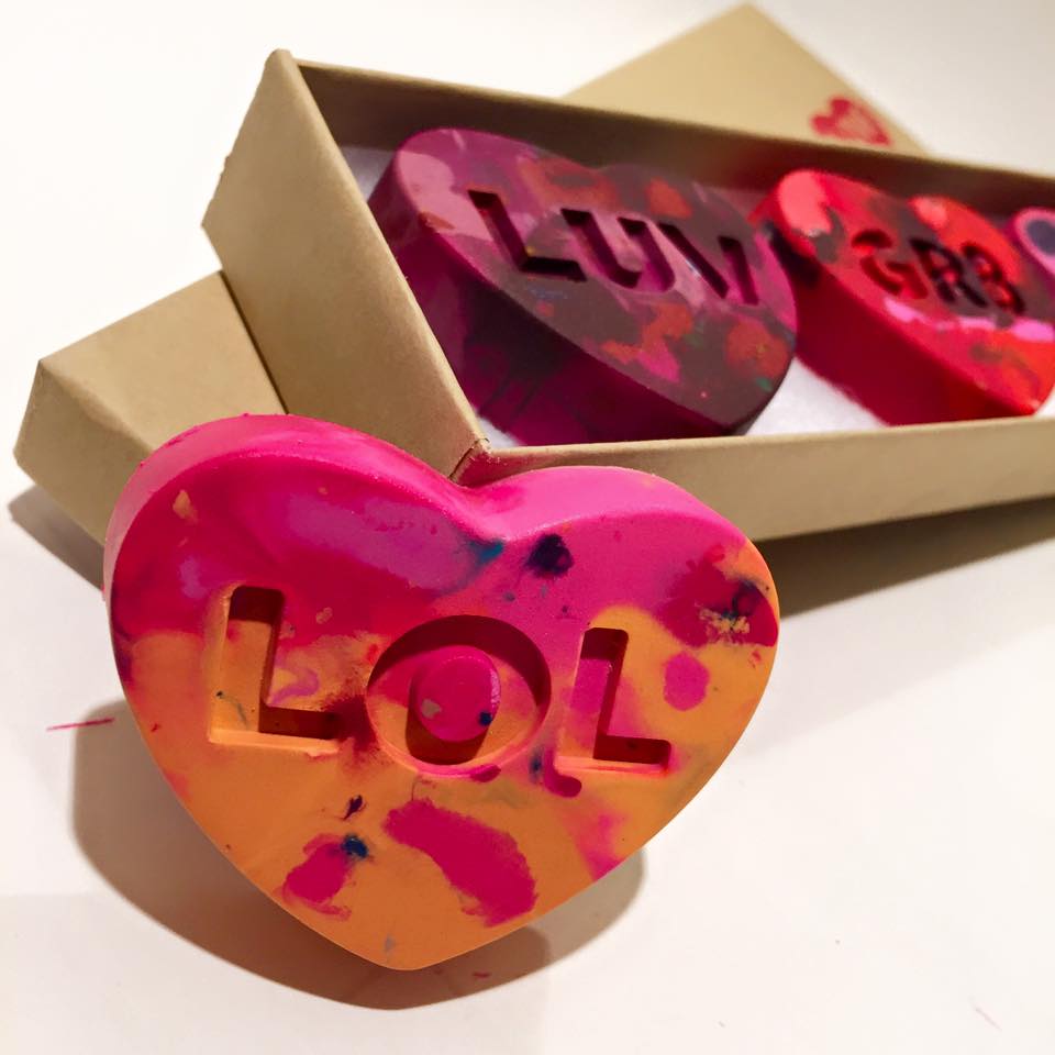 A single conversation heart rainbow crayon gift is propped up in front of a boxed set of three heart crayons. The crayon in the front says LOL and is made of mostly pinks and orange crayon colors. The box in the background is a kraft brown color and is displayed on a white background. 