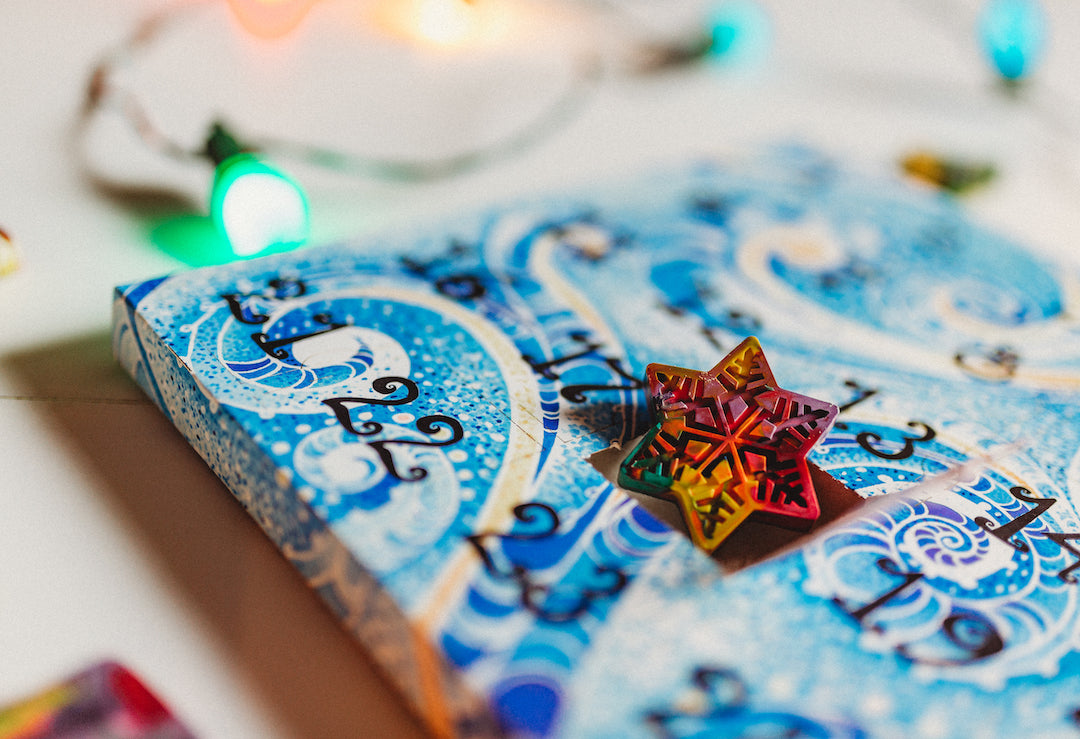 A blue crayon Advent calendar for kids sits on a white background with Christmas lights out of focus in the background. In box number 18, a mini Snowflake Original Rainbow Crayon is revealed with the paper calendar door open. Advent calendar is with a blue swirl design with black numbering on each of the punch out Advent Calendar doors. 