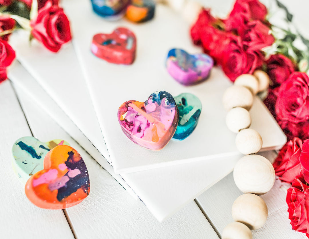 Closeup of 6 rainbow colored heart crayons with multicolors and swirls sits upon a white background with red roses around as a photo prop. 