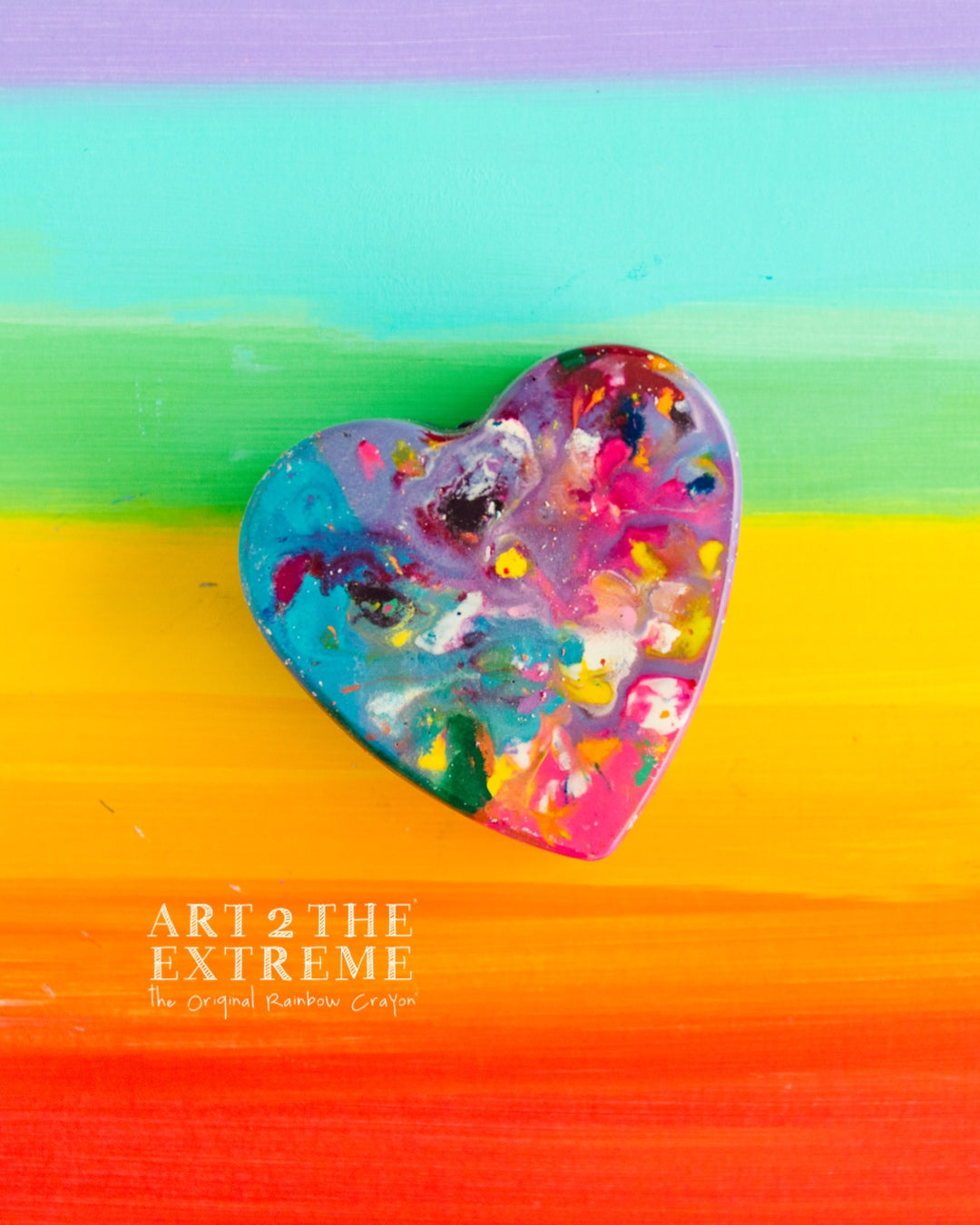 Multicolored, single, heart shaped jumbo crayon sits on top of rainbow paper that has purple on top fading down to red with teal, green, yellow, gold, orange in between as a photo prop background. This melted heart crayon almost looks tie dye and the purpose is to purchase to use as a special Valentine's Day crayon from handmade crayon shop, Art 2 the Extreme. 
