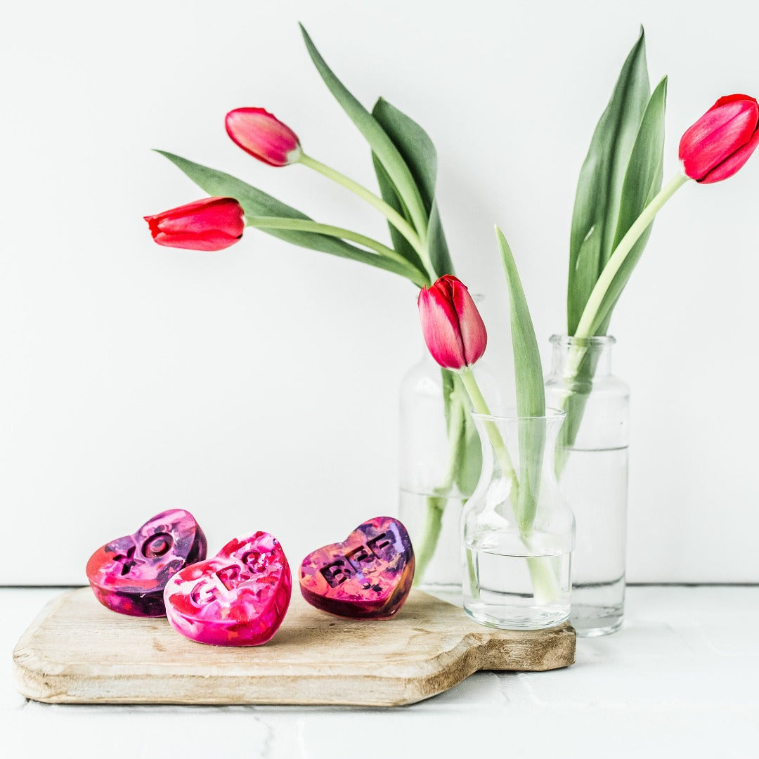 Three conversation heart crayons in pink/purple/red/peach combo.  Crayons are propped on a charcuterie board as a photo prop with a vase of red roses next to them.  The table and background are white. Multicolored heart crayons from Art 2 the Extreme make cute class valentines! 