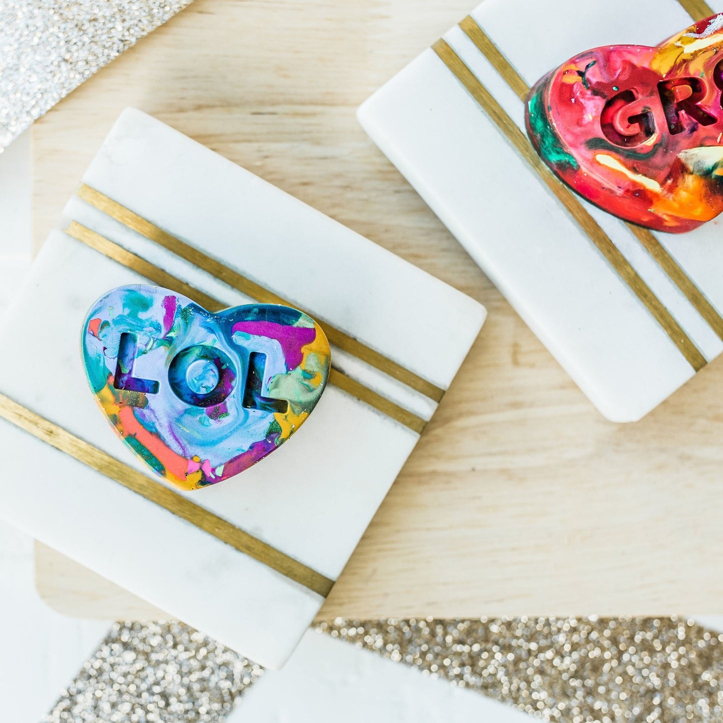 Multicolored heart crayon that has the letters LOL engraved. Each heart crayon is about 2 inches wide and made up of a variety of colors. This LOL crayon is mostly made of blues and teals. The crayon valentine is on a white coaster photoprop with a white and brown background. 