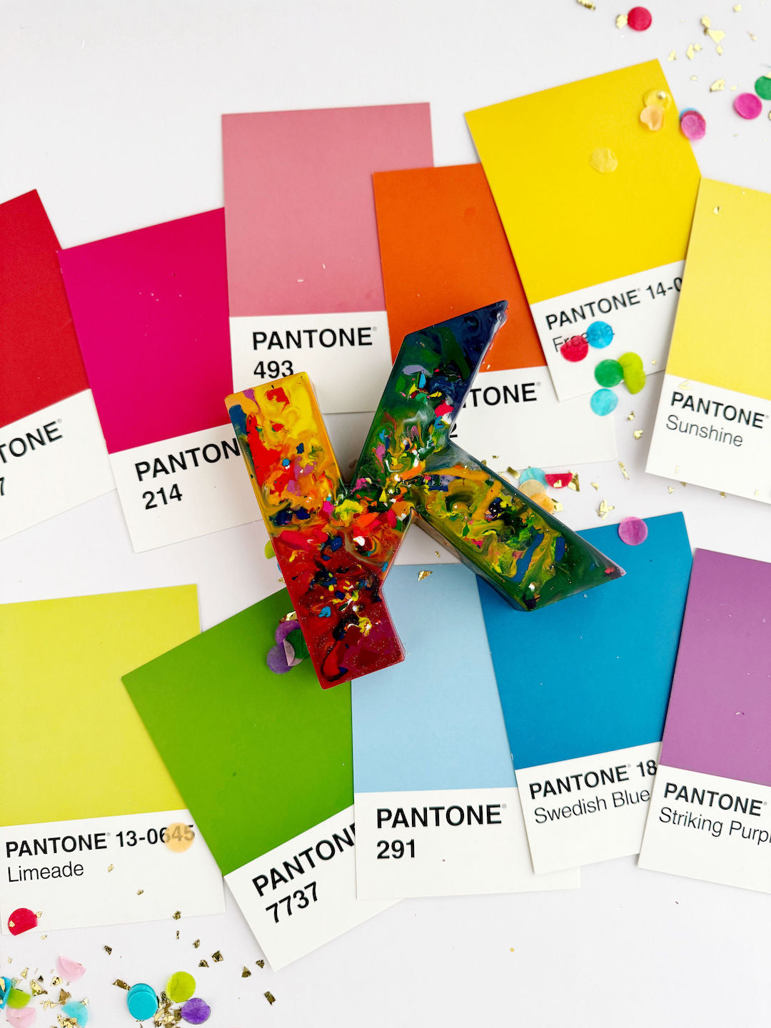 Large letter K oversized rainbow crayon in red, yellow, blue, and green with confetti like rainbow colors mixed in the crayon. Rainbow Crayon sits on Pantone color cards on a white background. 
