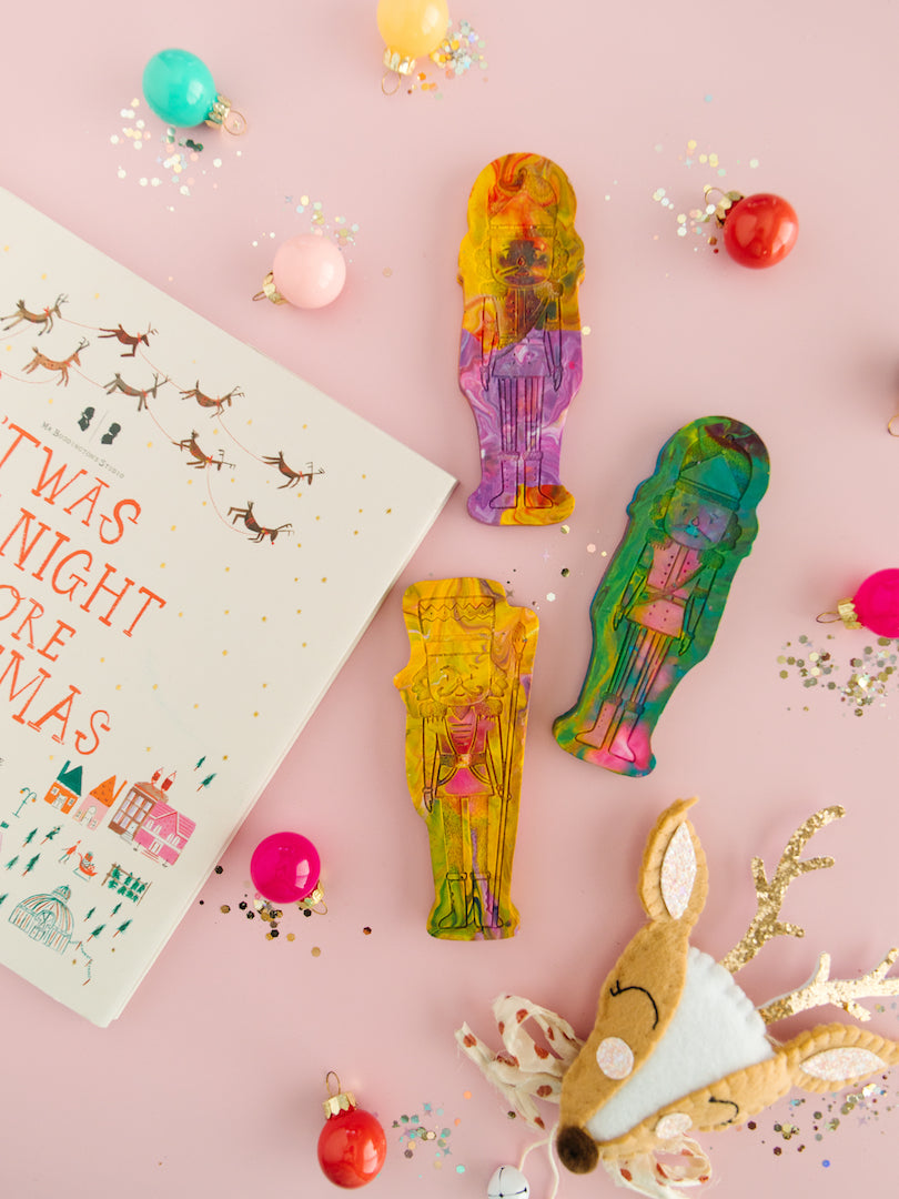 Three nutcracker ballet gifts crayons from Art 2 the Extreme on a pink background. These rainbow crayons are shaped like a classic nutcracker figure with vibrant multicolors swirled throughout. The crayon features intricate detailing, including a tall hat, mustache, and uniform-like design, making it a festive and creative holiday gift for children who love coloring. There is a Twas the Night Before Christmas book, ornaments, and glitter around the crayons as photo props for a festive photo. 