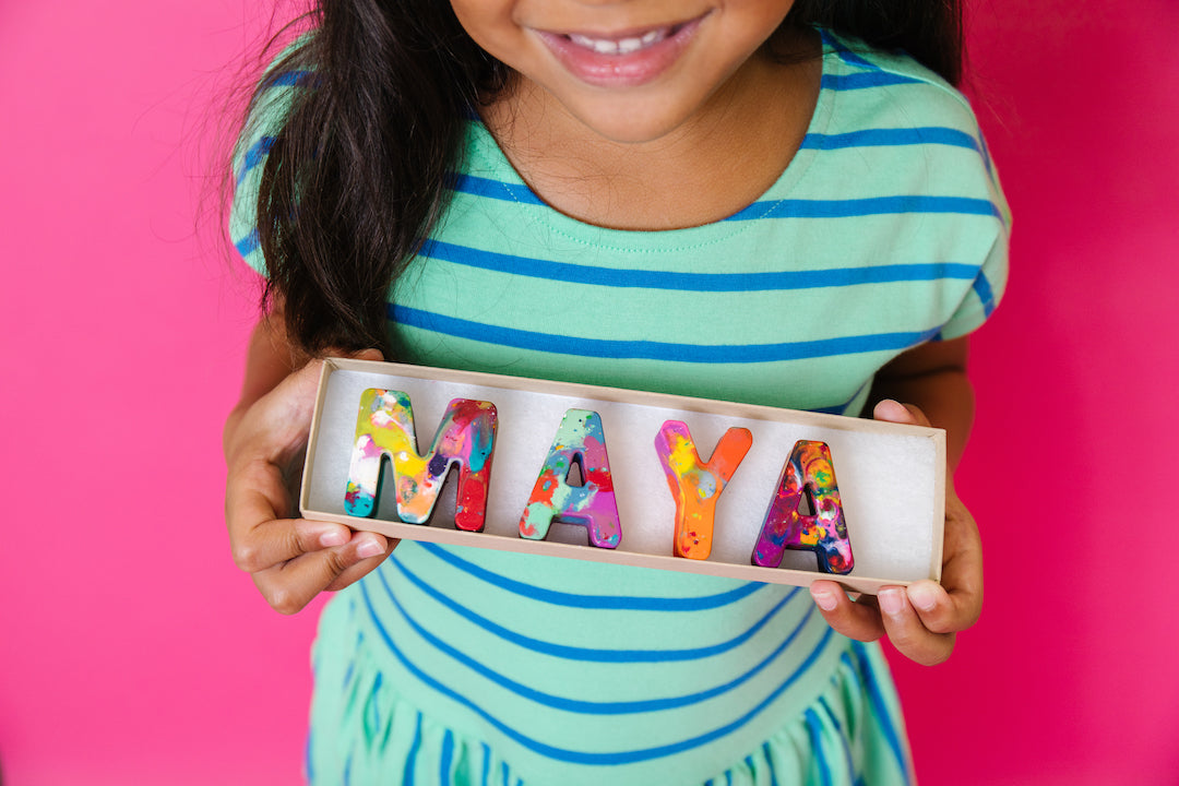 Girl holding a personalized name crayon set that spells MAYA in  rainbow crayon colors. The background is pink. This customized crayon gift is a perfect easter gift for little girls.  originalrainbow crayon.com
