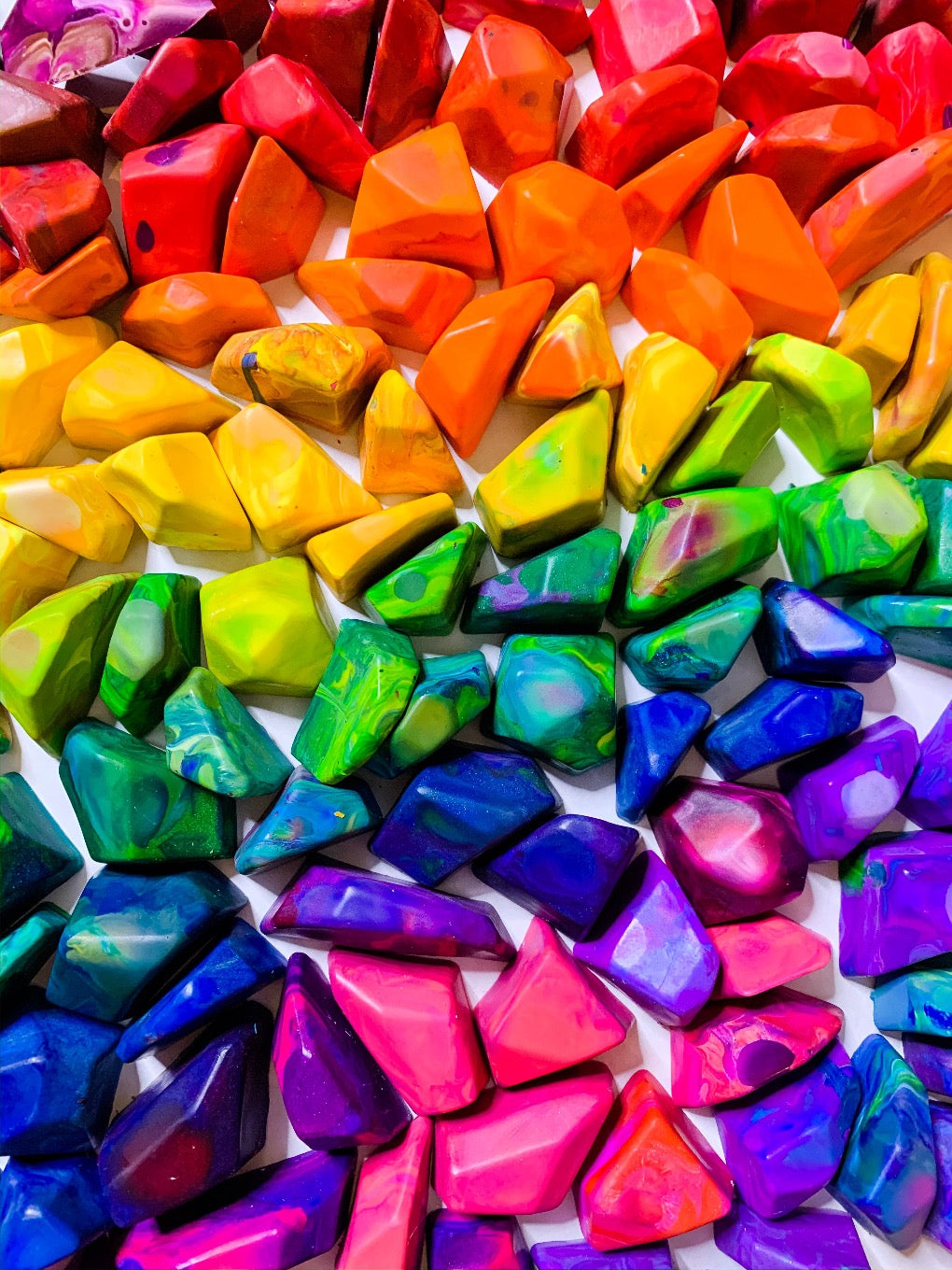 A ROY G BIV rainbow order display of multicolored crayon rocks from Art 2 the Extreme. Each crayon is in a multi-colored color family and will color in various colors. Top left of the image starts with the warmer hues such a pink, red, and orange. The middle has crayon rocks in the colors of yellow, green, then blue. The bottom is blue crayons, pink crayons, and purple crayon rocks. 