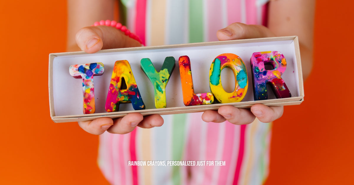 Child holding a boxed set of colorful personalized rainbow letter crayons spelling “TAYLOR,” with text reading “Rainbow Crayons, Personalized Just for Them.”