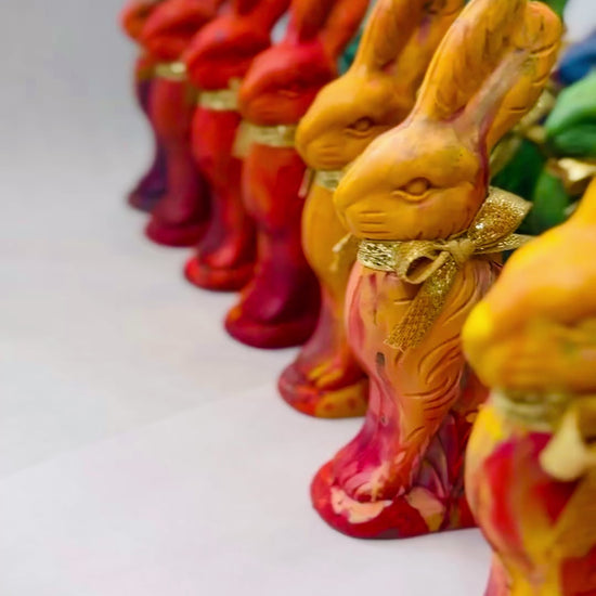 A colorful assortment of Easter Bunny crayons for kids, handcrafted in a rainbow of vibrant hues, each tied with a gold ribbon. A hand holding a brown Easter bunny crayon that looks like chocolate, but colors. Hand is turning bunny crayon with ears down to show off the crayons that color in multiple rainbow colors. Displayed in neat rows, the multicolored crayons offer a fun and creative alternative to traditional Easter treats and are unique Easter gifts for Easter.