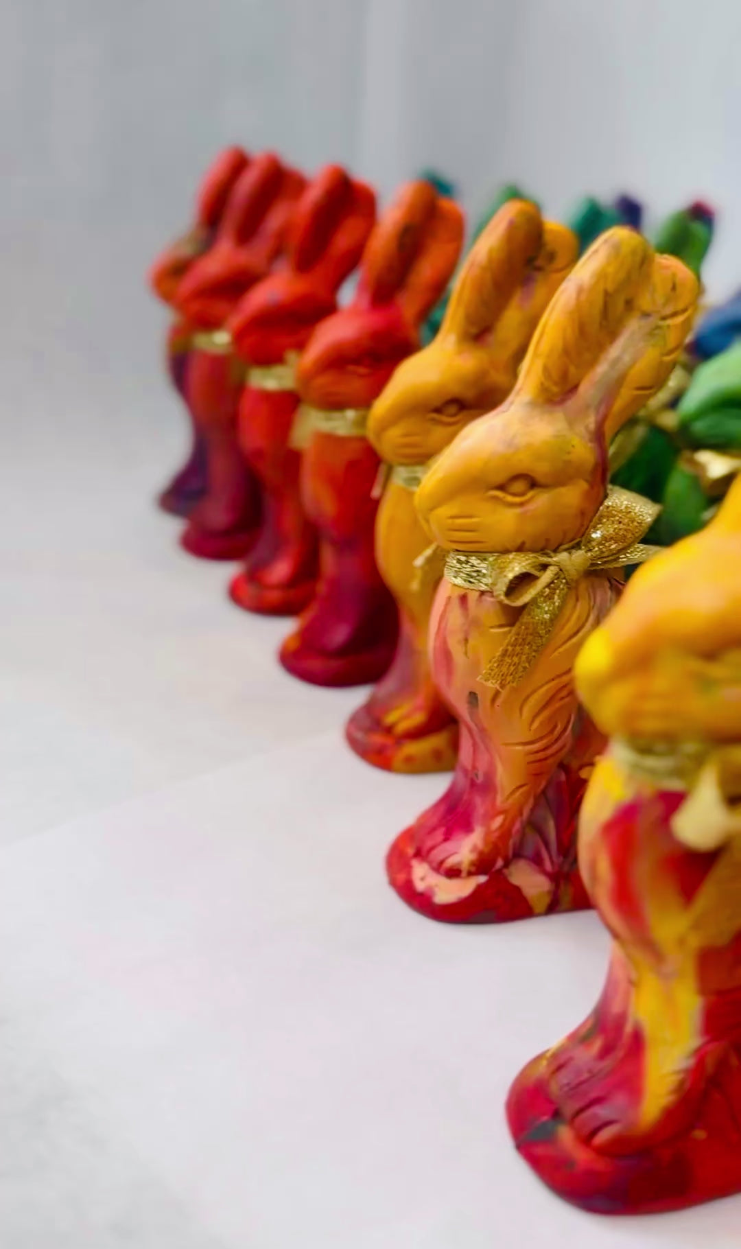 A colorful assortment of Easter Bunny crayons for kids, handcrafted in a rainbow of vibrant hues, each tied with a gold ribbon. A hand holding a brown Easter bunny crayon that looks like chocolate, but colors. Hand is turning bunny crayon with ears down to show off the crayons that color in multiple rainbow colors. Displayed in neat rows, the multicolored crayons offer a fun and creative alternative to traditional Easter treats and are unique Easter gifts for Easter.
